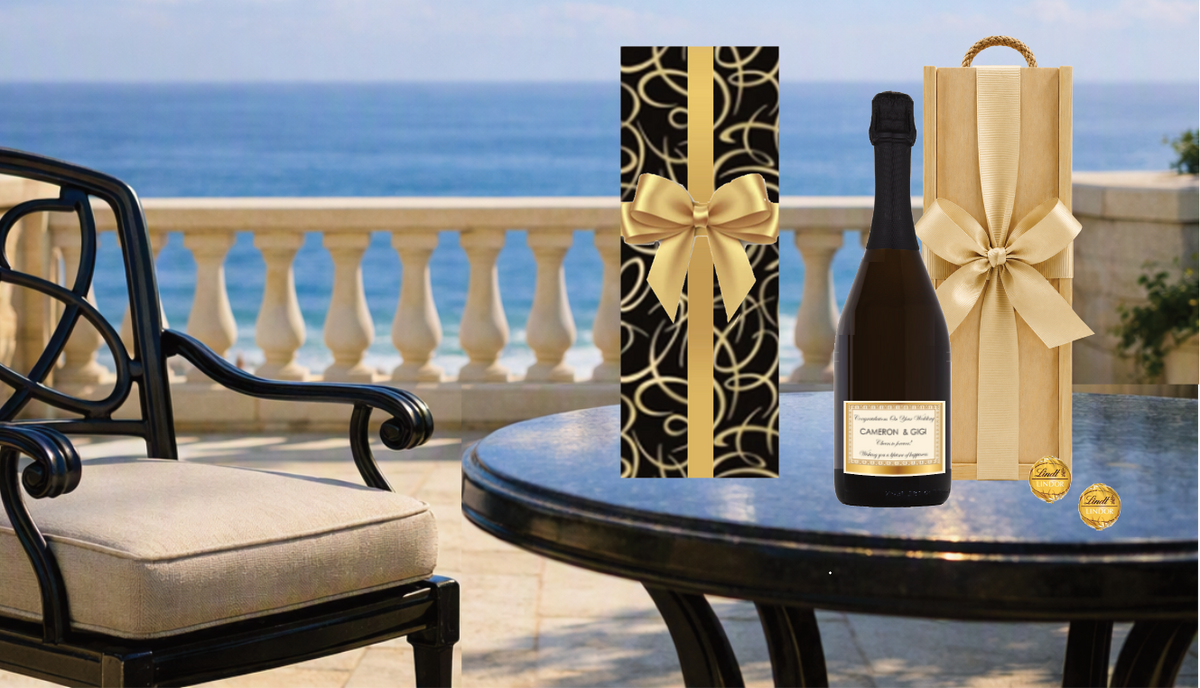 Prosecco & Truffles Gift box with personalised label . The gift is ready wrapped featuring gold swirls and a gold ribbon, set on a balcony table with an ocean view and a chair in the background.